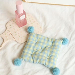 May include: A square, quilted cushion with a blue and yellow gingham pattern and four turquoise pom-poms at each corner. A pink perfume bottle and a floral-patterned hand mirror are also in the image.