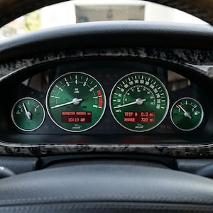 May include: A Jaguar vehicle instrument cluster featuring two large circular gauges with green faces, a tachometer on the left, and a speedometer on the right. Two smaller gauges indicate engine temperature and fuel level. Digital displays show 000001 mi.