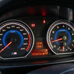 May include: A car dashboard featuring illuminated gauges. The speedometer has a blue and red design with the letter "M". The odometer displays 177,242 km. The time is 17:50 and the temperature is +9.0°C. The car is in park.