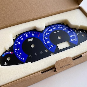 May include: A black and blue automotive instrument cluster gauge face overlay featuring white markings, the text RS, and KM/H, resting inside a custom-fitted foam insert within a brown cardboard box with the text Pull to Open on the flap.