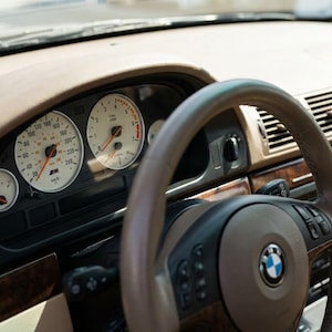 Könnte beinhalten: Innenansicht eines klassischen BMW-Autos. Das Armaturenbrett ist beige mit Holzverkleidung und Anzeigen mit weißen Zifferblättern und orangefarbenen Nadeln. Das Lenkrad ist braun und schwarz mit dem BMW-Logo.