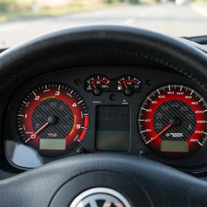 Pode incluir: Close-up do painel do carro com volante em couro preto. Os medidores têm detalhes em vermelho e preto, com números e ponteiros brancos. O velocímetro lê até 260. O carro está em uma estrada.
