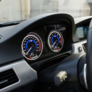 Può includere: Primo piano del cruscotto di un'auto con due indicatori illuminati. Gli indicatori hanno una combinazione di colori blu e rosso con segni bianchi. Il tachimetro mostra una velocità massima di 260 km/h. Il cruscotto è nero con finiture argentate.