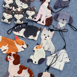 May include: A collection of dog-shaped accessories, including keychains and charms. The designs feature various breeds in cartoonish styles, with a variety of colors such as brown, white, black, and gray. Each accessory has a black lanyard attached.
