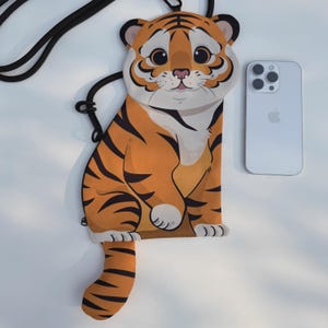 May include: A cartoon-style orange and black tiger-shaped crossbody phone bag with a black strap. The bag has a white belly and face, and a tail. A silver smartphone is next to the bag.
