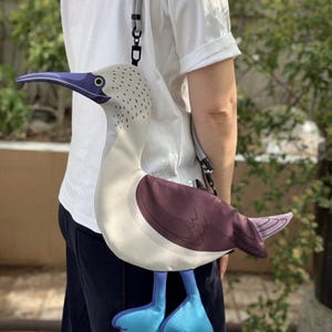 May include: A novelty crossbody bag shaped like a blue-footed booby bird. The bag features a white body, a purple back, and light blue feet. It has a long gray strap and a black clasp. The bird's head is white with a purple beak and a yellow eye.