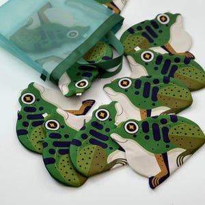 May include: A collection of frog-shaped coasters in shades of green, white, and brown. The coasters feature a detailed frog design with dark blue accents and a yellow and brown eye. A green mesh bag with a matching frog design is also present.