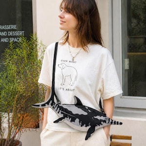 May include: A white t-shirt with the text "OVER WHAT" and a dog illustration. A black and white fish-shaped crossbody bag is worn across the body. The bag has a long strap and is made of a soft material.