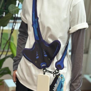 May include: A black and blue stingray-shaped crossbody bag with a matching strap. The bag has a white phone case attached to the bottom. The strap features blue fish and a black and white polka dot pattern.