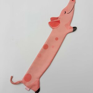 May include: A whimsical, long, pink pig-shaped bookmark with darker pink spots and accents. The pig has a smiling face, a curled tail, and black hooves. The bookmark is designed to be placed in a book.