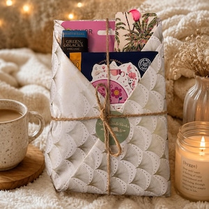 May include: A wrapped gift with a gold fan pattern, tied with twine. The gift contains books, a bookmark, and a small box of Green & Black's Organic chocolate. A lit candle and a mug of coffee are also in the image.