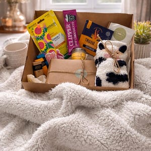May include: A gift box filled with self-care items, including a face mask, hot chocolate, coffee, a candle, and a pair of cow-print socks. The box is presented on a fluffy white blanket, with a mug and a vase of flowers in the background.