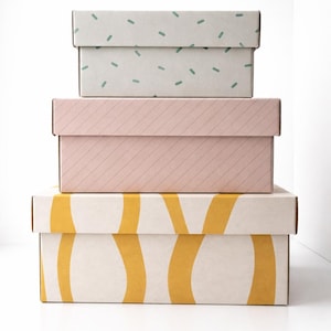 May include: Three stacked rectangular boxes with lids. The top box is light grey with small green dashes. The middle box is pink with diagonal lines. The bottom box is cream with yellow curved lines. All boxes are on a white background.