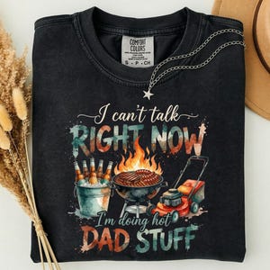 Può includere: Una t-shirt nera a girocollo con una grafica che riporta il testo I can't talk RIGHT NOW I'm doing hot DAD STUFF. Il design include un secchiello di birre, un barbecue a carbone con fiamme e un tosaerba rosso su uno sfondo ad acquerello.