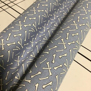 May include: A roll of fabric featuring a repeating pattern of off-white bones scattered across a light blue background. The material is tightly wound, showing a consistent print of various bone shapes oriented in multiple directions across the surface.