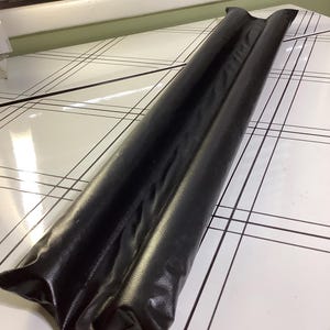 May include: A long, black door draft stopper made of a shiny, leather-like material. The stopper is rectangular and has a folded design. It is placed on a white surface with a black grid pattern.