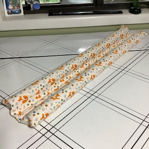 May include: A long, cylindrical fabric draft stopper featuring a repeating pattern of orange foxes, brown acorns, and small blue and green leaves on a cream-colored background. The item is constructed from two parallel tubes of stuffed fabric.