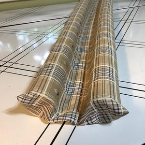 May include: A long, cylindrical fabric draft stopper featuring a tan, white, and black plaid pattern with small, scattered white floral accents. The textile has a visible seam running along the top and is finished with gathered fabric at the ends.