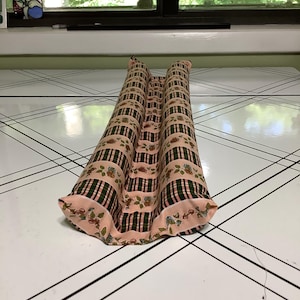 May include: A long, cylindrical fabric draft stopper featuring a repeating pattern of dark green and black plaid stripes interspersed with bands of small, light pink floral motifs on a pale peach background, with gathered fabric ends.