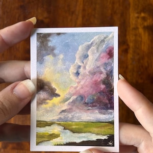 May include: A rectangular art print featuring a landscape painting of a marshland under a dramatic sky. The scene displays a winding waterway reflecting light, green grasses, and large, textured clouds in shades of white, pink, and dark blue.
