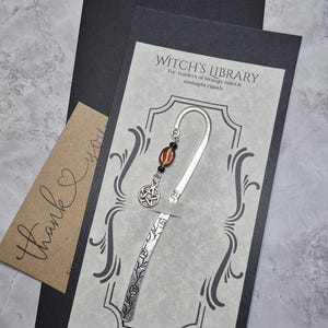 Puede incluir: Un marcapáginas plateado con detalles florales y un dije de pentagrama, presentado en una tarjeta con el texto "Witch's Library". El marcapáginas tiene una cuenta decorativa y se presenta con una tarjeta de "gracias".