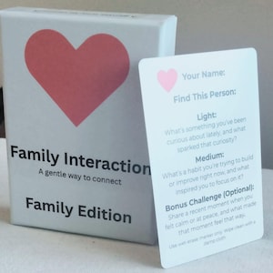 May include: A white box with a large red heart and the words "Family Interaction" and "Family Edition" in black. A card with a pink heart and prompts for self-reflection is next to the box.