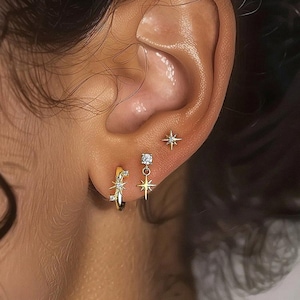 May include: Close-up of an ear adorned with three gold earrings. One is a small hoop with a starburst design, another is a dangling star, and the third is a small star stud. All earrings have small, clear gemstones.