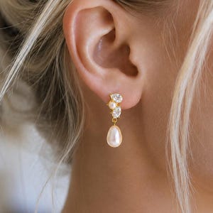May include: A gold-tone drop earring featuring a cluster of two clear pear-shaped crystals and one small round white pearl at the post, with a single larger teardrop-shaped white pearl suspended from the base of the crystal cluster.