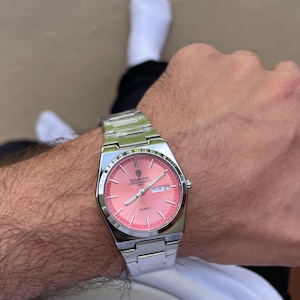 Könnte beinhalten: Eine silberfarbene Quarzuhr aus Edelstahl mit einem runden, lachsfarbenen Zifferblatt. Das Zifferblatt hat silberne Stundenmarkierungen, silberne Zeiger und eine Datumsanzeige auf der Drei-Uhr-Position. Der Text BINBOND FASHION WATCH 1853 ist aufgedruckt.