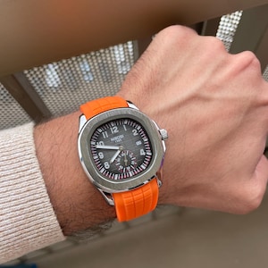 Peut inclure: Une montre-bracelet avec un boîtier carré argenté et un cadran noir texturé doté de chiffres arabes blancs, d'aiguilles argentées et d'un petit sous-cadran. La montre possède un bracelet en caoutchouc orange vif avec un motif quadrillé et le texte NIBOSI 1985.