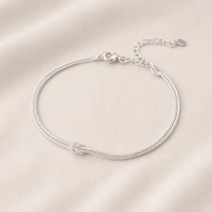 Könnte beinhalten: Ein silberfarbenes Kettenarmband mit einem zarten Knotendetail in der Mitte. Das Armband besteht aus einer feinen, flexiblen Schlangenkette und verfügt über eine verstellbare Verlängerungskette mit einem kleinen, flachen, herzförmigen Anhänger.