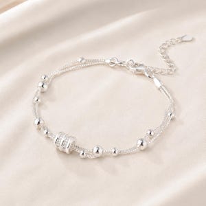 May include: A double-strand silver bracelet featuring a central cylindrical bead encrusted with small clear stones, interspersed with smooth silver spheres of varying sizes along the delicate chain, finished with a lobster clasp and extender chain.