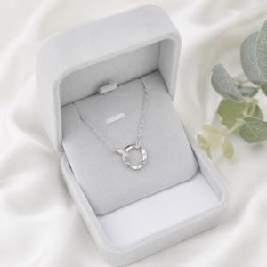 May include: A silver-toned necklace featuring a delicate chain and a circular pendant with a hammered, textured finish. The pendant is suspended from the chain, which is displayed inside a light gray, velvet-lined jewelry box with a hinged lid.