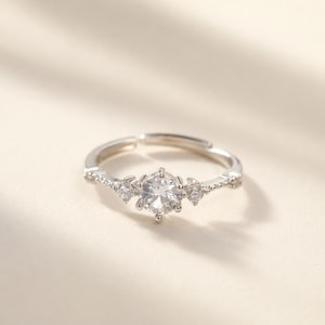 Könnte beinhalten: Ein silberfarbener, verstellbarer Ring mit einem zentralen, rund geschliffenen, klaren Stein, der auf jeder Seite von zwei kleineren klaren Steinen flankiert wird. Die Ringschiene ist dünn und metallisch mit kleinen eingebetteten Steinen.