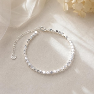 Op de afbeelding: Een zilverkleurige armband bestaande uit kleine, gefacetteerde, kubusvormige kralen in een doorlopend cirkelvormig patroon. De armband heeft een fijn verlengkettinkje met een kleine, ronde, gegraveerde bedel aan het uiteinde van de sluiting.