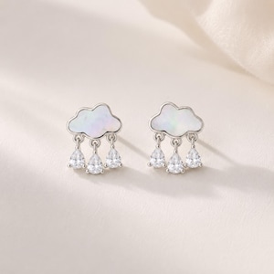 May include: A pair of silver stud earrings shaped like clouds with iridescent white inlay. Each cloud features three dangling, pear-shaped clear crystals that resemble raindrops, suspended from small silver loops attached to the base of the cloud.