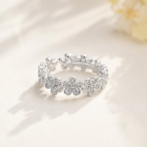 May include: A silver-toned ring featuring a continuous band of interconnected flower shapes. Each individual flower motif is encrusted with small, clear, sparkling stones that catch the light, creating a delicate and uniform circular design.