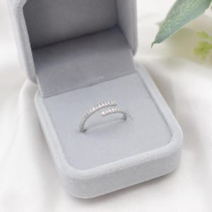 Puede incluir: Un anillo de estilo bypass en tono plateado con un diseño abierto y dos extremos superpuestos, cada uno adornado con una hilera de pequeñas piedras claras y brillantes, presentado dentro de un joyero forrado en terciopelo gris claro.