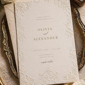Może przedstawiać: Kremowe zaproszenie ślubne z tłoczonymi kwiatowymi obwódkami i złotym napisem. Tekst brzmi: Kindly join us for the wedding of Olivia and Alexander, August 20th at 5 PM, 123 Anywhere St., Any City, ST 12345, reception to follow.