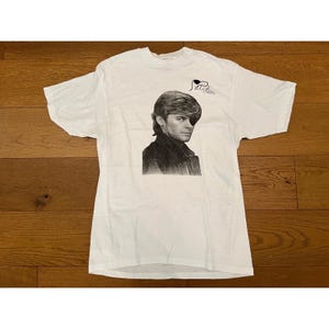 May include: White t-shirt featuring a black and white portrait of a man with an eye patch. The shirt has the word "Patch" written above the portrait in a cursive font. The t-shirt is made of a soft, comfortable material.