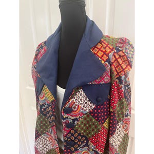 May include: A blazer featuring a patchwork design with various geometric, floral, and paisley patterns in red, green, blue, and yellow. The garment includes a solid navy blue collar, matching navy blue pocket flaps, and three navy blue buttons.