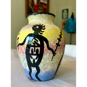 May include: A ceramic vase with a rounded body and a narrow neck, featuring a multi-colored sponge-painted background in yellow, pink, and blue. A large, black, stylized figure with outstretched arms is painted on the front surface.