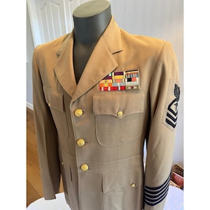 Puede incluir: Una chaqueta de uniforme militar color canela con botones dorados, dos bolsillos en el pecho y una hilera de cintas multicolores en el lado izquierdo. La manga muestra una insignia de rango negra bordada y cuatro rayas horizontales en el puño.