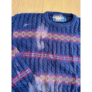 Könnte beinhalten: Ein dunkelblauer Zopfmuster-Pullover mit einem horizontalen Muster aus violetten, orangefarbenen und türkisfarbenen Rauten. Am Kragen befindet sich ein Etikett mit dem Text Ralph Lauren und drei Bärenabbildungen. Der Stoff zeigt helle Verfärbungen.