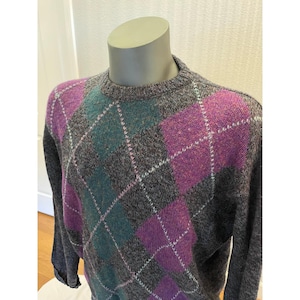 Könnte beinhalten: Ein langärmeliger Pullover mit Rundhalsausschnitt und einer dunkelgrauen, melierten Basis mit einem markanten Argyle-Muster. Die Rauten sind in leuchtendem Lila und Türkis gehalten und mit feinen, hellen Linien durchzogen.