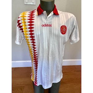 May include: A white short-sleeve polo shirt with a red collar and a vertical pattern of red and yellow diamonds on the right side. The shirt features a red adidas logo on the chest and a red and yellow Spanish national crest on the left side.