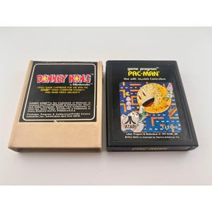 Pode incluir: Dois cartuchos de videogame retangulares para o sistema Atari. O cartucho esquerdo é bege com um rótulo preto escrito Donkey Kong by Nintendo. O cartucho direito é preto com um rótulo colorido apresentando o Pac-Man e o logotipo Atari.