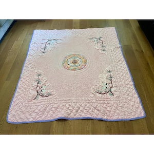 May include: A light pink quilted blanket featuring a central circular medallion with floral and crane motifs. The corners are decorated with embroidered branches and white cranes. The blanket has a textured surface and a light purple border.