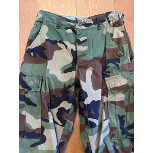 Puede incluir: Pantalones cargo con estampado de camuflaje boscoso en tonos verde oliva, marrón y negro. Los pantalones incluyen dos grandes bolsillos laterales tipo cargo con solapas, cierre de botón en la cintura y una hebilla metálica de ajuste lateral.