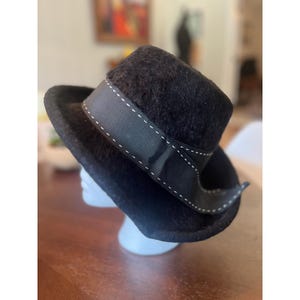 May include: A black fuzzy textured hat with a wide brim and a rounded crown. The hat features a black ribbon band wrapped around the base, detailed with a single row of white decorative stitching along the top and bottom edges of the ribbon.
